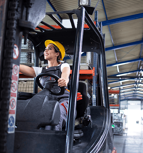 Woman wearing a yellow hard hat seated at the controls of a forklift in a warehouse