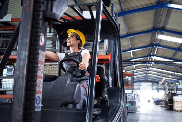 Woman wearing a yellow hard hat seated at the controls of a forklift in a warehouse