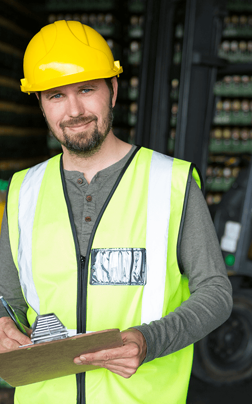 Warehouse worker wearing a hard hat and high-visibility vest holding a clipboard beside a forklift