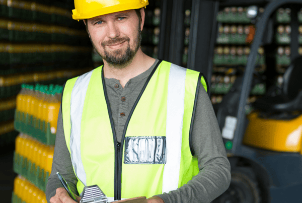 Warehouse worker wearing a hard hat and high-visibility vest holding a clipboard beside a forklift