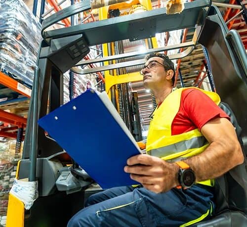 Forklift operator holding a blue clipboard while seated in a warehouse forklift