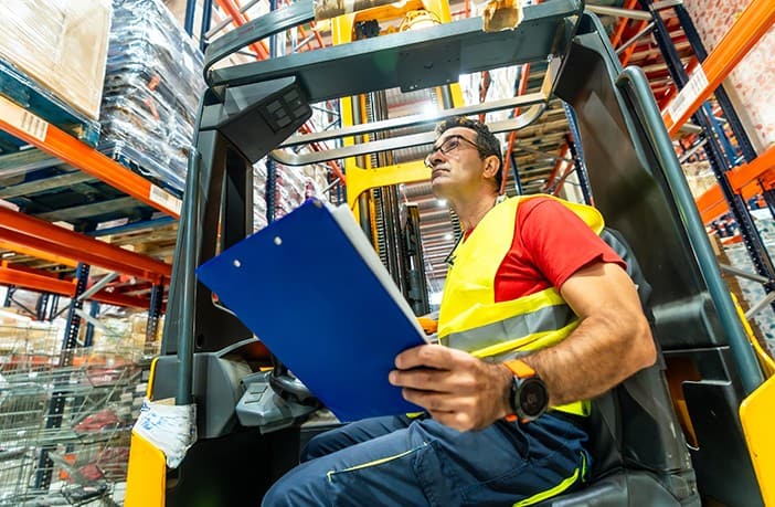 Forklift operator holding a blue clipboard while seated in a warehouse forklift